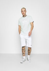 Gaiši piparmētru zaļš kokvilnas T-krekls ar "ALPHA INDUSTRIES" logo, apvienots ar baltām šortiem. Pelēki sporta apavi, redzamas tetovējumi uz rokām un kājām.