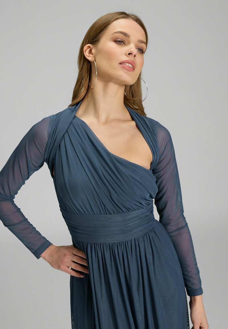 Sininen verhoiltu mekko, jossa on pitkät läpikuultavat hihat, istuva yläosa ja liehuva helma. Mekossa on olkaimeton muotoilu ja poimutettu vyötäröyksityiskohta.