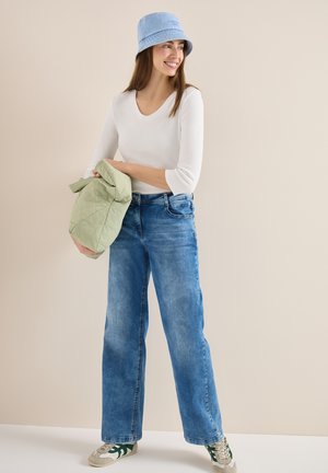 Frau trägt blauen Anglerhut, weißes Langarmshirt, weit geschnittene Jeans und Turnschuhe und hält eine hellgrüne gesteppte Jacke.