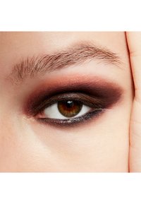 Nahaufnahme eines Auges mit dunklem, rauchigem Lidschatten, der in satte Brauntöne übergeht. Die Iris ist braun mit einem Hauch von Grün, umgeben von natürlichen Wimpern.