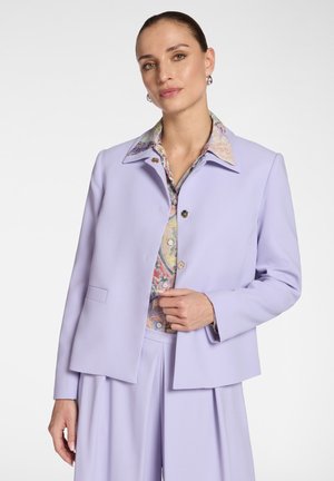 Femme portant un blazer violet clair et un pantalon plissé assorti sur une chemise à motifs colorés, debout devant un fond blanc uni.