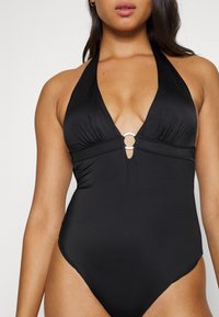 Costum de baie negru, dintr-o singură piesă, cu decolteu halter adânc, având un accent central sub formă de inel, material adunat și deschideri mari pe picioare.