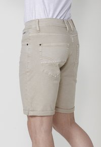 Man med beige jeansshorts med uppvikta fållar och en vit t-shirt, sedd bakifrån mot en enkel vit bakgrund.