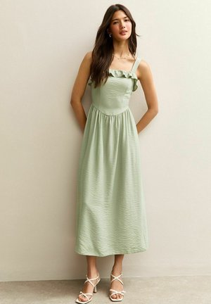 Vestido maxi verde claro con tirantes con volantes, tela texturizada, cuerpo ajustado y cintura fruncida; combinado con sandalias blancas de tacón y tiras.