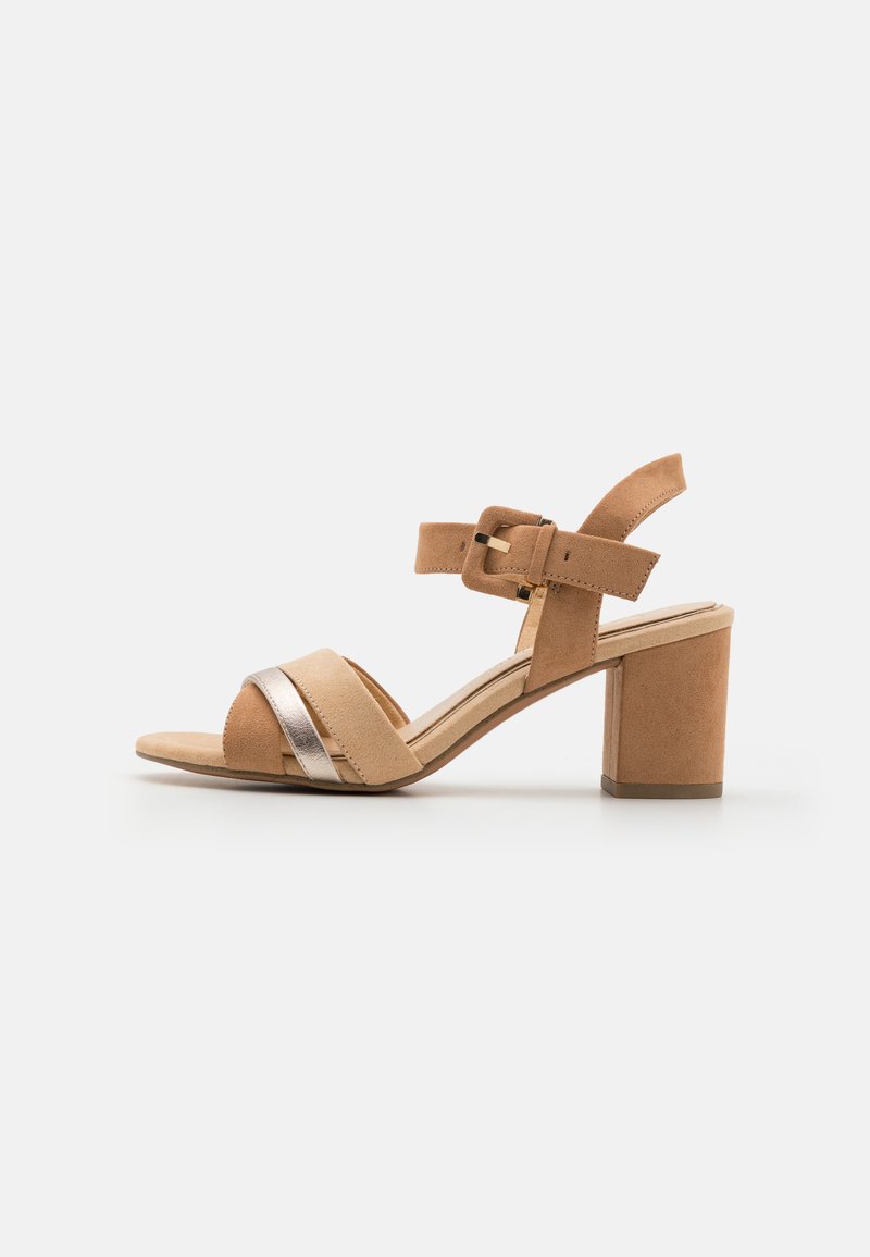 Tan suede heeled sandal with a metallic strap, ankle buckle, and block heel. Open-toe design with soft texture and neutral color palette.
