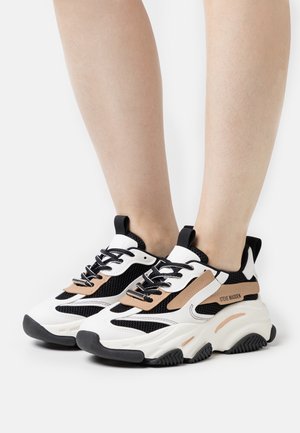 Pieds portant des baskets épaisses Steve Madden noires, blanches et beiges avec des détails en mesh et cuir sur un fond blanc.