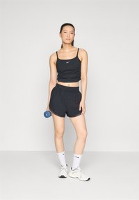 Čierny športový crop top s tenkými ramienkami a malým logom, kombinovaný s čiernymi šortkami, bielymi ponožkami a bielymi teniskami, drží modrý činkový závaží.