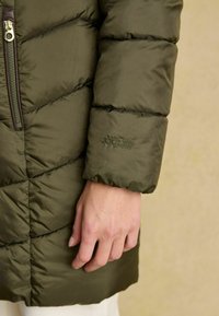 Olivovo zelená puffra bunda s diagonálnym šitím a zipsovou kapsou; na manžete je vyšívané logo. Hladká, ľahká tkanina.