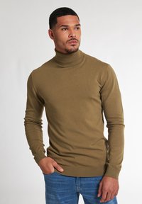 Hnědý rolák ze měkké, elastické pleteniny. Má dlouhé rukávy a přiléhavý střih, kombinovaný s modrými džíny pro kontrast.