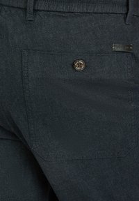 Marineblauwe textuur broek met een achterzak met een donkere knop en een klein label op de tailleband, benadrukt een gladde stofafwerking.