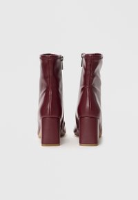 Stivaletti alla caviglia in pelle ecologica bordeaux con un design aderente, dotati di tacco a blocco e una cerniera laterale per una facile vestibilità. Texture liscia in tutto.