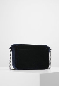 Tmavě modrá semišová kabelka přes rameno s klopou a stříbrným řetízkovým popruhem. Hladká textura a obdélníkový tvar s zaoblenými hranami.