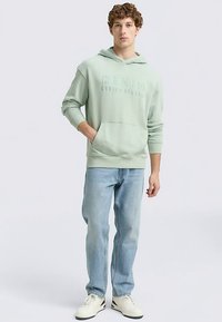 Šviesiai žalias džemperis su kapišonu ir priekiniu kišeniu bei reljefiniu užrašu, derinamas su šviesiai mėlynais džinsais ir baltais sportbačiais. Minkšta, kasdienė tekstūra.