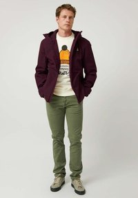 Chaqueta impermeable marrón sobre un suéter blanco con un gráfico de sombrero naranja, pantalones verdes y zapatillas beige con suelas negras.