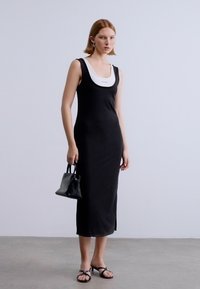 Μαύρο αμάνικο φόρεμα με εφαρμοστή σχεδίαση, με λευκό εσωτερικό γύρο λαιμού. Το μοντέλο κρατά μια μικρή μαύρη τσάντα χειρός και φοράει μαύρα σανδάλια.