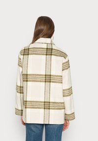 Rutig jacka i kräm och olivgrön, med krage, långa ärmar och en avslappnad passform. Mjuk textur med distinkta rutmönster.