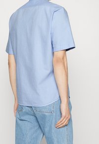 Hombre vistiendo una camisa de manga corta azul clara y jeans azul claros, mostrado desde atrás contra un fondo liso claro.