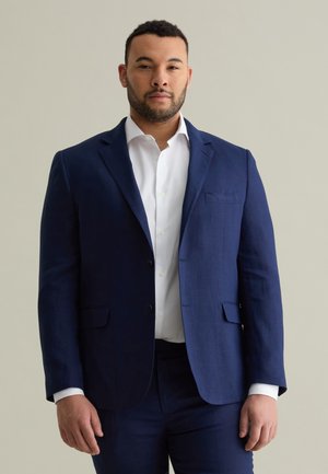 Veste de costume bleu marine et pantalon assorti avec une chemise blanche. La veste a un col cranté, deux boutons et des poches latérales.