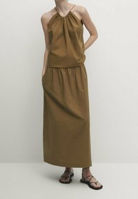 Robe marron sans manches avec encolure froncée, silhouette fluide et poches latérales ; portée avec des sandales plates noires. Design simple et uni.