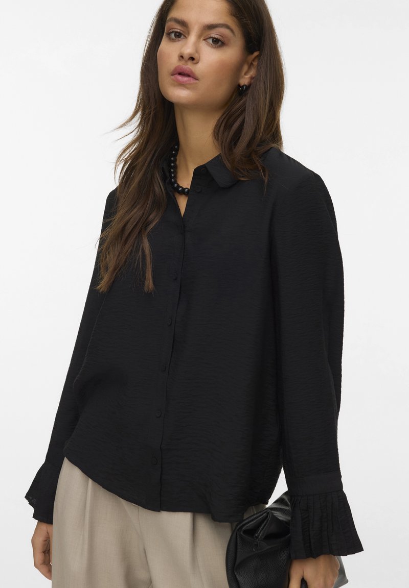 Black long-sleeve button-down shirt with textured fabric, featuring a relaxed fit and ruffled cuffs, paired with beige trousers and a black bag.