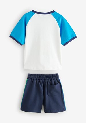 Bijela majica s kratkim rukavima i plavim raglan rukavima, u paru s mornarskim kratkim hlačama koje imaju zelenu bočnu prugu i elastičnu traku u struku.