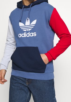 Hoodie en bloc de couleurs bleu, rouge et bleu marine. Il présente un grand logo Adidas blanc, des manches longues et une poche kangourou sur le devant. Mélange de coton.