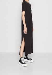 Czarna sukienka do kolan z krótkimi rękawami i bocznymi rozcięciami. Noszona z czarnymi sneakersami za kostkę z białą podeszwą. Prosty design, miękka faktura.