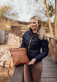 Braune Leder-Crossbody-Tasche mit strukturiertem Finish, ausgestattet mit zwei Reißverschlussfächern und einem geprägten Logo, gehalten von einer Person in einer schwarzen Jacke.