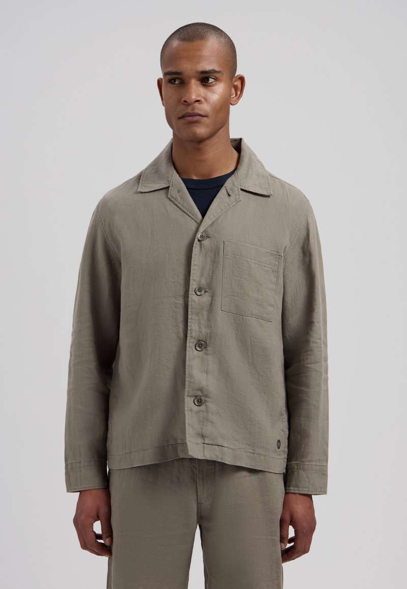 Homme portant une veste beige à manches longues boutonnée avec poche poitrine par-dessus une chemise noire, debout devant un fond gris uni.