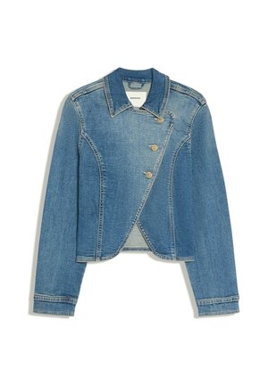 Kvinde med langt mørkt hår står udenfor en bygning iført en cropped denimjakke, vide cowboybukser og spidse brune sko.