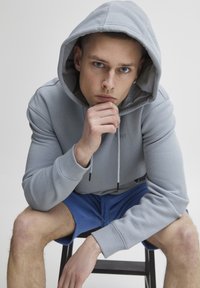 Lys grå hoodie i blødt stof med snoretræk i huen, parret med blå shorts. Model sidder på en stol, hånd på hagen, og viser en afslappet pose.