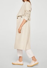 Abrigo largo beige con cuello doblado, frente abotonado doble y bolsillos laterales, combinado con pantalones blancos recortados y zapatillas claras.