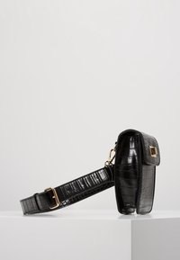 Svart krokodilprickad handväska med en strukturerad design, gyllene detaljer och en justerbar axelrem, med en lockstängning och texturerad yta.