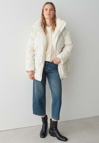 Giacca imbottita bianca con un colletto peloso, indossata sopra un cardigan beige e jeans larghi blu, abbinata a stivaletti neri alla caviglia.