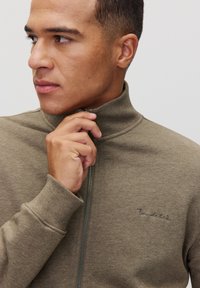 Sweatshirt zippé marron avec un col haut, texture tricotée et logo brodé. Modèle tenant la fermeture éclair, mettant en avant la coupe ajustée.