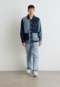 Jeansjakke med en blanding av mørke og lyse paneler, med to store frontlommer og knappefeste, paret med lyseblå jeans.