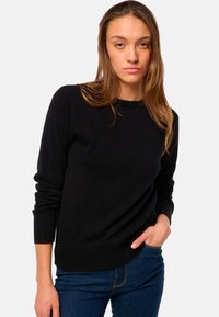 Pull en maille noire à manches longues, col rond, avec des poignets et un ourlet côtelés. Porté avec un jean bleu foncé, mettant en avant un look élégant et décontracté.