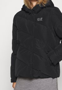 Chaqueta acolchada negra con diseño acolchado, con capucha y un logo de EA7. Los detalles incluyen bolsillos laterales y una textura suave y lisa.