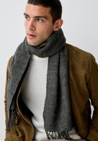 Écharpe à carreaux grise en laine douce, avec des bords frangés, enroulée autour du cou d'un modèle. Portée sur un pull blanc en maille et une veste marron.