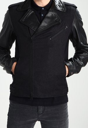 Personne portant une veste noire avec des manches et un col en cuir, mains dans les poches et jean foncé sur un fond uni.