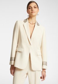 Blazer de couleur crème avec un col et des poignets embellis de métal, coupe structurée, fermeture à un bouton et deux poches avant.