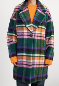 Kvinne iført en fargerik oversized rutete frakk med grønne, rosa, oransje, lilla og hvite mønstre over en oransje genser.