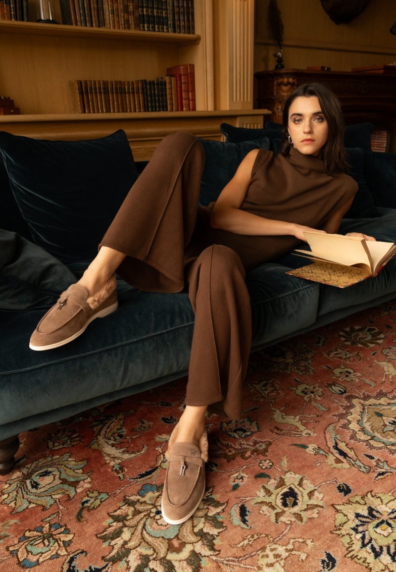 Bruna mockaläders loafers med pälsfoder och vit sula. Modell i matchande mörkbrun outfit, sittande på en mörkgrön sammetssoffa och läser.