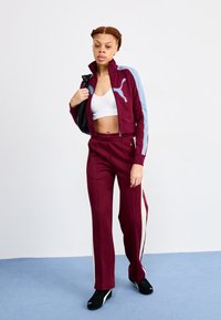 Bordeauxkleurige trainingspak met lichtblauwe accenten, bestaande uit een ritsjack en high-waisted broek. Het model houdt een zwarte tas vast en draagt een witte top.