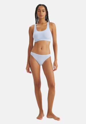 Kobieta z splecionymi włosami stojąca boso, ubrana w jasnoniebieski, bezszwowy biustonosz sportowy i pasujące bikini na białym tle.