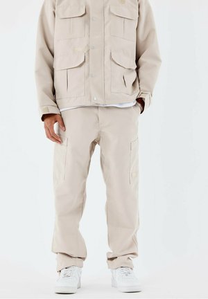 Mężczyzna w beżowych spodniach cargo i pasującej kurtce, ubrany w białe tenisówki, stoi na tle jednolitego białego tła.