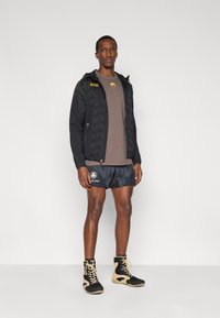 Μαύρο αθλητικό μπουφάν με γεωμετρικό σχέδιο, καφέ πουκάμισο, μαύρα σορτς με κυματιστό μοτίβο και μαύρα/χρυσά αθλητικά μποτάκια.