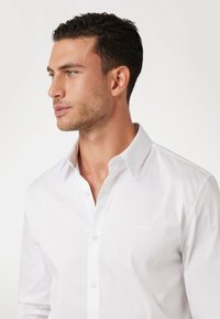 Camisa blanca de algodón con cuello clásico, textura sin escamas y logo bordado en el pecho. Silueta ajustada.