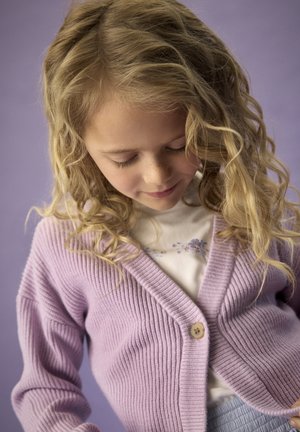 Lavendelfarbener, gerippter Cardigan mit V-Ausschnitt, versehen mit einem einzelnen Holzknopf, getragen über einem weißen Oberteil mit Blumenmuster. Welliges blondes Haar umrahmt das Gesicht.