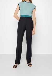 Szydełkowa turkusowa bluzka z krótkimi rękawami i czarnymi paskami, zestawiona z czarnymi spodniami o wysokim stanie i czarnymi sandałami na obcasie.